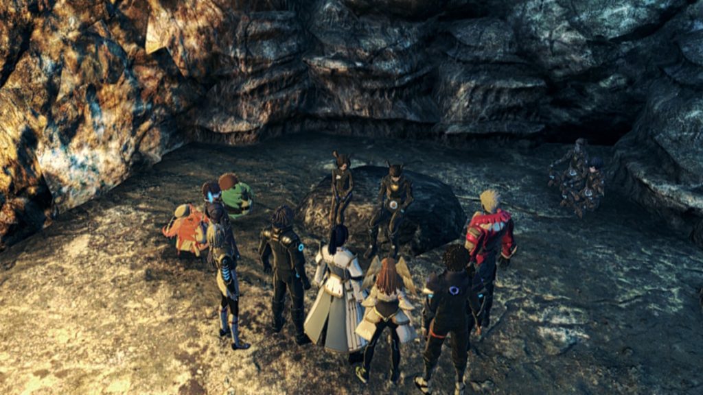 A group of character in a variety of different outfits stand in a semi-circle around two people in similar soldier garb sitting on a rock inside a cave